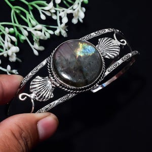 Op de afbeelding: Een zilverkleurige armband met een grote, ronde, donkergrijze labradoriet edelsteen met iriserende blauwe en gele flitsen. De metalen band is aan weerszijden van de centrale steen versierd met twee gedetailleerde schelpmotieven.