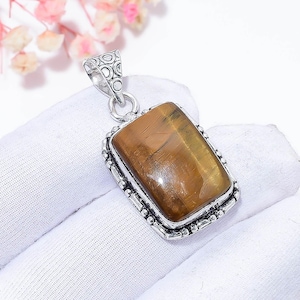 May include: A rectangular pendant featuring a polished, golden-brown tiger's eye stone. The stone is set in a decorative silver-colored metal frame with a detailed bail for a chain. The pendant is displayed on a white surface.