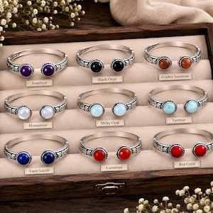Puede incluir: Una colección de nueve brazaletes abiertos de tono plateado dispuestos en una bandeja de madera. Cada brazalete presenta dos piedras preciosas redondas, etiquetadas como Amethyst, Black Onyx, Golden Sunstone, Moonstone, Milky Opal, Larimar, Lapis Lazuli, Carnelian y Red Coral.