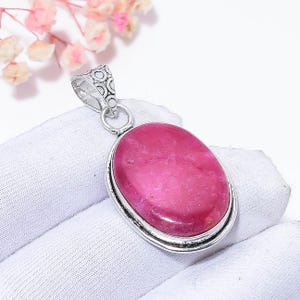 Puede incluir: Colgante de plata con una piedra preciosa rosa ovalada. La piedra está engastada en un bisel de plata y tiene una superficie pulida. El colgante tiene una fianza decorativa con un diseño circular. El fondo es blanco.