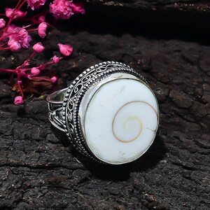 May include: A silver ring featuring a round, white Shiva eye shell with a natural spiral pattern. The setting is crafted with intricate, oxidized metal filigree detailing around the circular stone, creating a textured and ornate appearance.