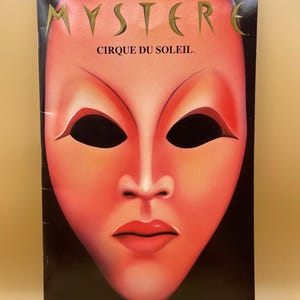 May include: Souvenir program for Mystère by Cirque du Soleil. The cover features a stylized red face with large black eye holes and red lips. The word "MYSTÈRE" is at the top, and "CIRQUE DU SOLEIL" is below it.