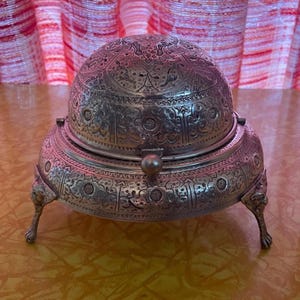 Puede incluir: Una mantequera antigua de color plateado con intrincados patrones florales y geométricos. El plato tiene una tapa abovedada y se apoya en tres patas ornamentadas con forma de garra. El diseño sugiere un artículo vintage o de colección.