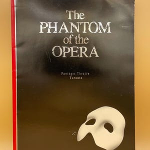 May include: A black theatre programme for "The Phantom of the Opera" with white text and a white mask graphic. The title is in large, silver letters. The spine has a red stripe. Location: Pantages Theatre, Toronto.