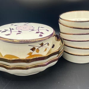 May include: A set of vintage ceramic serving dishes. The set includes a covered dish with a floral design and a stack of small bowls. The dishes have a cream-colored base with gold and purple trim.