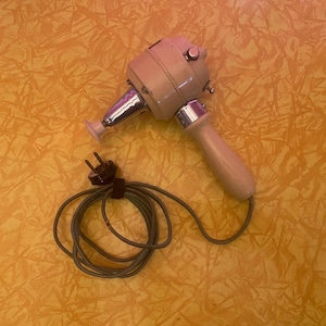 Puede incluir: Un masajeador de mano vintage beige con un cabezal plateado y un mango beige claro. El masajeador tiene un cable de alimentación con un enchufe negro. El cable está enrollado en espiral. El masajeador está sobre una superficie amarilla.