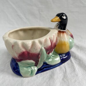 Può includere: Una fioriera a forma di anatra in ceramica con testa nera, becco giallo e corpo colorato. La fioriera ha un interno bianco ed è decorata con accenti rossi, verdi e blu. L'anatra è appollaiata su una base blu.