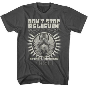 May include: Dark gray t-shirt with the text "DON'T STOP BELIEVIN' JOURNEY DETROIT, MICHIGAN 1980" in a distressed, vintage style. The design features a circular emblem with wings and a sunburst pattern.