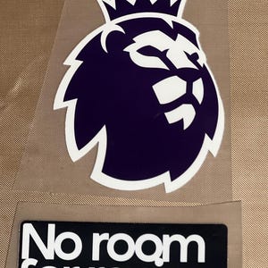May include: A purple and white lion logo with a crown, and the text "No room for racism" on a black rectangle. The lion logo is a stylized design, and the text is in a bold, sans-serif font.