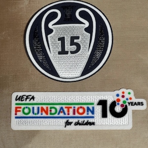 May include: Two patches. The top patch is a navy blue and white circular design with a silver trophy and the number 15. The bottom patch is rectangular with the text "UEFA FOUNDATION for children" and "10 YEARS".