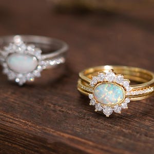 Könnte beinhalten: Zwei Ringe mit ovalen Opal-Edelsteinen. Ein Ring ist silberfarben mit einem Kranz aus kleinen, klaren Steinen. Der andere Ring ist goldfarben, ebenfalls mit einem Kranz. Die Ringe liegen auf einer dunklen Holzoberfläche.