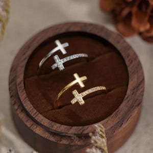 May include: Two open-style rings featuring cross motifs, one in silver-tone metal and one in gold-tone metal. Each ring has a plain cross on one end and a cross encrusted with small clear stones on the other, resting inside a round wooden box.