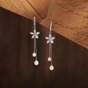 Op de afbeelding: Een paar zilverkleurige oorbellen met een vijfbladige bloemvorm ingelegd met heldere stenen. Elke oorbel heeft twee fijne kettinkjes die aan de bloem hangen, met aan het uiteinde van elke ketting een kleine witte parel.