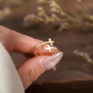 May include: A gold-toned open ring featuring two cross motifs. One cross is embellished with small, clear, sparkling stones along its surface, while the other cross is plain, polished gold. The ring has a slender, adjustable band design.