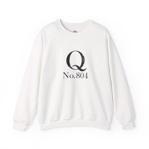 May include: A white crew neck sweatshirt with long sleeves. The front displays a large black letter "Q" above the text "No.804". The sweatshirt is made of a soft material, suitable for casual wear.
