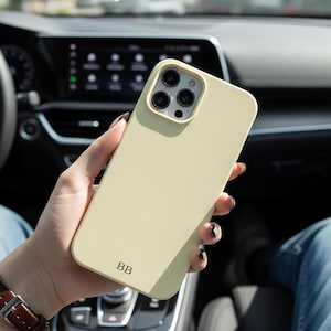 Puede incluir: Funda de teléfono amarillo claro con recorte para la cámara y las iniciales "BB". La funda se sostiene en una mano, con el interior de un coche en el fondo. El color es amarillo pálido.
