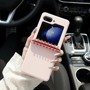 May include: A pale pink phone case with a notepad-style design. The case features a camera aperture and a screen showing the time. The notepad section has a red spiral binding. The phone case is held in a car.