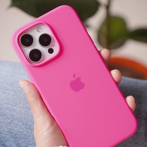 Puede incluir: Una funda de teléfono rosa brillante con el logotipo de Apple. La funda tiene un acabado liso y está diseñada para un teléfono inteligente con múltiples lentes de cámara. La funda del teléfono está en la mano de una persona.