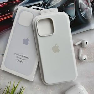 Könnte beinhalten: Eine weiße Silikonhülle für das iPhone 15 Pro Max mit Apple-Logo, neben der Verpackung. Auf der Verpackung steht "iPhone 15 Pro Max Silicone Case". Weiße kabellose Ohrhörer und ihr Ladecase sind ebenfalls abgebildet.
