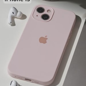May include: A pale pink iPhone 13 with a matching case and a pair of white wireless earbuds. The phone case has a matte finish and features the Apple logo. The text "iPhone 13" is in the upper left corner.
