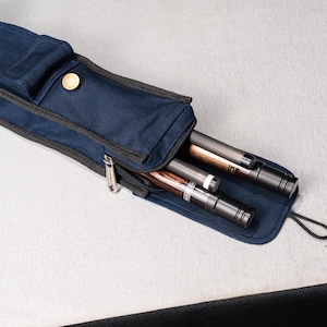 Op de afbeelding: Een marineblauwe keu-etui met een ritssluiting en een gouden knoopsluiting. De etui is open en onthult drie keuen met zwarte en bruine accenten. De etui heeft een zwarte koordlus.