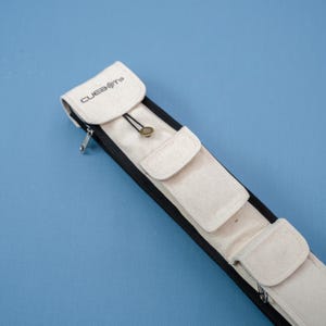 Op de afbeelding: Een beige biljartkeuikoffer met de tekst "CUEBOTS" op de flap. De koffer heeft drie kleine vakken en een zwarte rits. De koffer staat op een blauwe achtergrond.