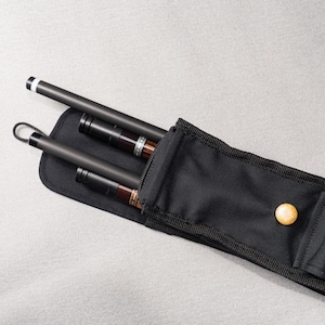 Op de afbeelding: Een zwarte draagtas met drie donkere poolkeuen. De keuen hebben bruine en zilveren accenten. De tas heeft een drukknoopsluiting en een klein vakje. De achtergrond is lichtgrijs.