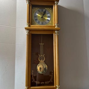 May include: A wooden grandfather clock with a gold-colored clock face and pendulum. The clock has Roman numerals and decorative gold accents. The pendulum is a large, gold-colored sphere.