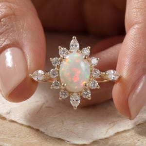 Könnte beinhalten: Ein Goldring mit einem zentralen ovalen Opal, der von einem Kranz aus kleinen, klaren, marquise- und rundgeschliffenen Steinen umgeben ist. Die Fassung besteht aus einem polierten Goldband, das eine strahlenförmige Anordnung bildet.