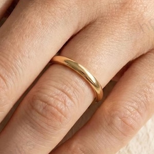 Könnte beinhalten: Ein goldfarbener Ring mit einem glatten, abgerundeten Band wird an einem Finger getragen. Der Ring hat ein schlichtes, elegantes Design. Der Ring ist wahrscheinlich aus Gold oder einem goldfarbenen Metall gefertigt. Der Ring ist ein Schmuckstück.