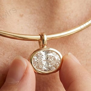 Könnte beinhalten: Goldfarbene Halskette mit einem ovalen Diamantanhänger. Der Anhänger wird von einer Hand gehalten, wodurch das Design der Halskette hervorgehoben wird. Die Halskette ist im Choker-Stil gehalten.