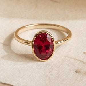 May include: A gold ring featuring a single oval-cut deep red gemstone set in a smooth bezel setting. The band is thin and polished, showcasing a minimalist design that highlights the vibrant colour and clarity of the central red stone.