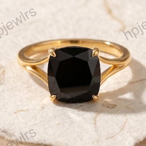 Könnte beinhalten: Ein goldener Ring mit einem großen, quadratischen, schwarzen Edelstein. Der Ring liegt auf einer strukturierten, cremefarbenen Oberfläche. Das Ringband hat ein schlichtes, elegantes Design.