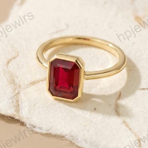 Könnte beinhalten: Ein goldener Ring mit einem rechteckigen, tiefroten Edelstein. Der Ring liegt auf einer strukturierten, cremefarbenen Oberfläche, mit dem Markennamen "hpjewlrs" im Hintergrund. Das Design des Rings ist schlicht und elegant.