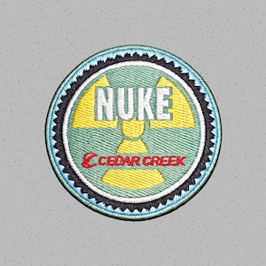 Könnte beinhalten: Runder, gestickter Aufnäher mit dem Wort "NUKE" in Weiß, einem gelben Strahlungssymbol und dem Text "CEDAR CREEK" in Rot. Der Aufnäher hat einen tealfarbenen Hintergrund mit schwarzen und blauen Rändern.