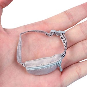 Puede incluir: Una pequeña riñonera gris claro con cremallera plateada y una correa con la palabra "GENIC" en blanco. La correa tiene una hebilla de plástico gris. La riñonera está diseñada para llevarse alrededor de la cintura.