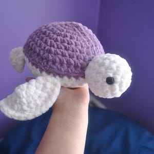 May include: A handmade crochet turtle in shades of purple and white. The turtle has a textured, rounded shell in a light purple color, with white accents on the head, flippers, and belly. The turtle has a black eye.