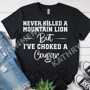 Puede incluir: Camiseta negra con el texto blanco "NEVER KILLED A MOUNTAIN LION But I'VE CHOKED A Cougar". La camiseta se muestra con vaqueros azules y zapatillas negras y blancas. También se ve una pequeña planta suculenta.