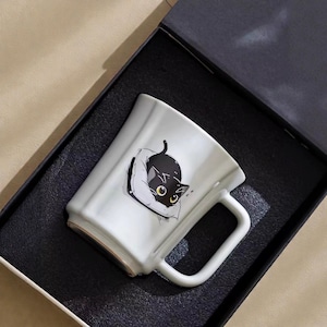 Może przedstawiać: Biały ceramiczny kubek z ilustracją czarnego kota. Kot leży na poduszce, z żółtymi oczami i czarnym ciałem. Kubek ma kwadratowy kształt i uchwyt. Prezentowany w czarnym pudełku prezentowym.