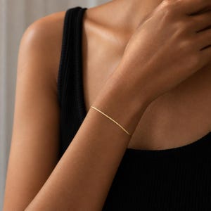 May include: A thin, delicate gold chain bracelet worn around the wrist. The bracelet features a smooth, flat, and reflective surface that catches the light, creating a subtle metallic sheen against the skin. The design is simple, slender, and refined.