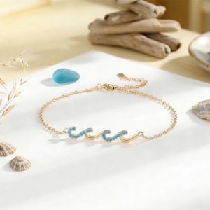 May include: A gold-colored bracelet with a wave design, adorned with small, light blue gemstones. The bracelet is displayed on a white surface, alongside seashells and decorative elements. The bracelet is a delicate piece of jewelry.