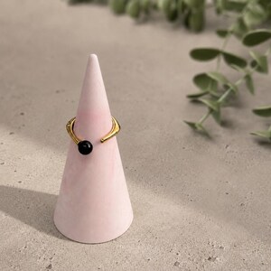 Op de afbeelding: Een goudkleurige ring met een zwarte kraal wordt getoond op een roze, kegelvormige ringhouder. De ringhouder staat op een lichtgrijze ondergrond, met groen gebladerte op de achtergrond. De ring heeft een open ontwerp.