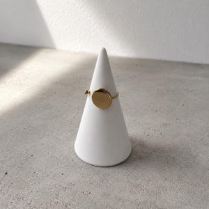Op de afbeelding: Een goudkleurige ring met een plat, cirkelvormig schijfelement en een klein bolvormig detail op de ringband. De ring wordt gepresenteerd op een witte, kegelvormige standaard en toont de gepolijste metalen afwerking en het open ontwerp.