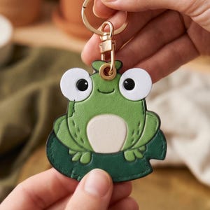 Könnte beinhalten: Ein froschförmiger Schlüsselanhänger aus Leder in Grün mit großen weißen Augen, schwarzen Pupillen, einem hellbeigen Bauch und einer dunkelgrünen Seerosenblatt-Basis. Der Anhänger ist an einem goldfarbenen Ring mit Karabiner befestigt.