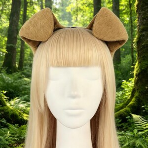 May include: A headband featuring two plush, tan-colored dog ears. The ears are attached to a black headband, designed to be worn on the head. The background is a blurred forest scene.