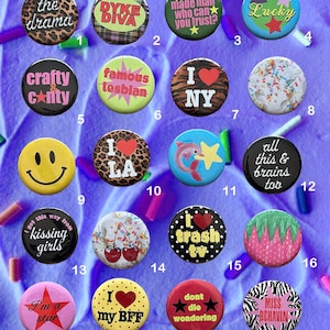 May include: A collection of twenty colorful pinback buttons with various designs and text. The buttons feature phrases like "Lucky," "I Love NY," and "I'm a star." The buttons are round and have a safety pin backing.