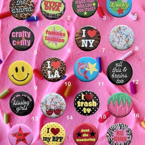 May include: A collection of twenty colorful pinback buttons with various designs and text, including phrases like "DYKE DIVA," "Lucky," and "I love NY." The buttons are arranged on a pink surface with sprinkles.