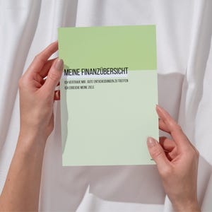 Puede incluir: Una tarjeta verde claro y blanca sostenida por dos manos. La tarjeta tiene el texto "MEINE FINANZÜBERSICHT" en la parte superior, con texto adicional debajo. El fondo es una tela blanca texturizada.