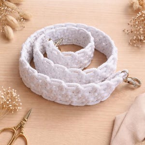 以下が含まれることがあります： 銀色の留め金が付いた、手作りの白いマクラメバッグストラップ。 ストラップは柔らかい織り素材でできており、明るい木製の表面に巻かれています。 上部には「Handmade MACRAME BAG STRAP」の文字があります。