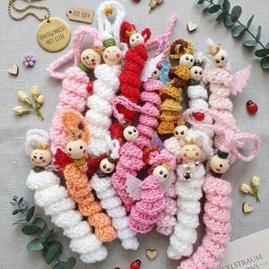 May include: A collection of handmade crochet dolls in various colors, including pink, white, and orange. Each doll has a wooden head with a painted face and is adorned with small details like wings and crowns. The dolls are arranged with decorative elements.
